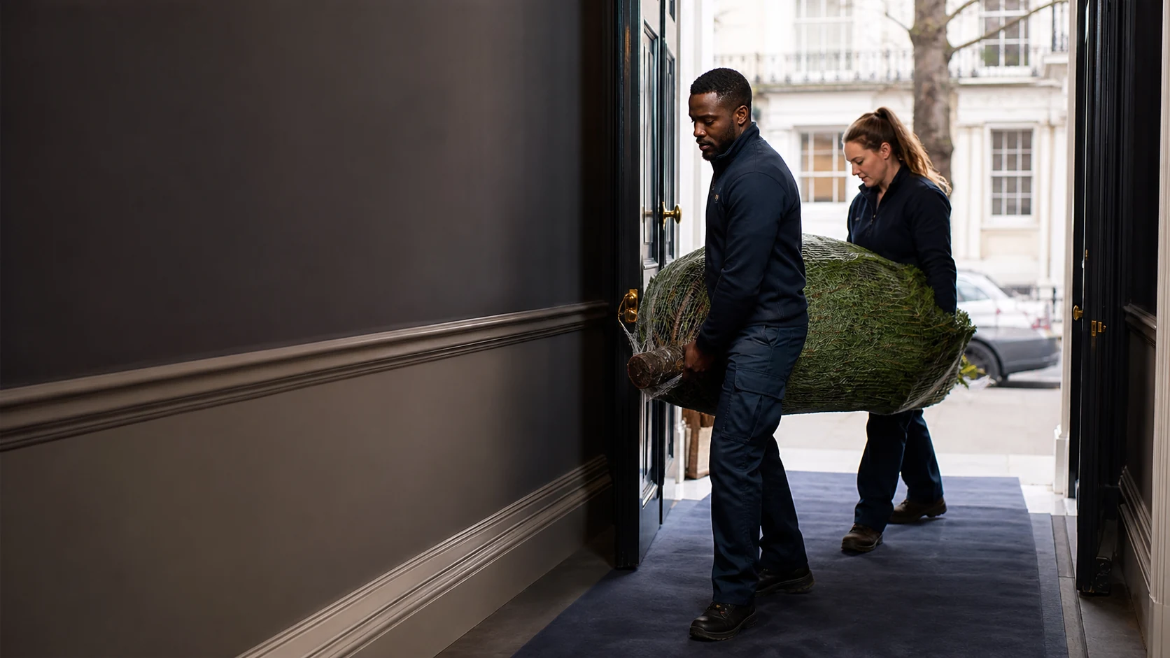 Delivery professional carrying a wrapped real Christmas tree toward a London townhouse